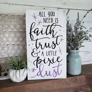 May include: White wooden sign with the text "All you need is faith, trust & a little pixie dust" in black lettering with purple glitter accents.