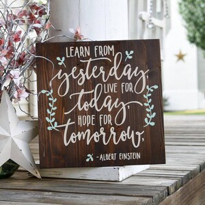 May include: A wooden sign with a dark brown stain and a white painted quote: "Learn from yesterday, live for today, hope for tomorrow." The quote is attributed to Albert Einstein. The sign has a light blue painted floral design.