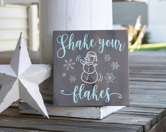May include: A square dark brown wooden sign featuring the light blue text "Shake your flakes" and a white line-art illustration of a snowman surrounded by several white snowflake designs, positioned next to a white five-pointed star ornament.