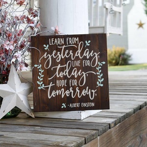 May include: A wooden sign with a dark brown stain and a white painted quote: "Learn from yesterday, live for today, hope for tomorrow." The quote is attributed to Albert Einstein. The sign has a rustic look and is decorated with green leaves.