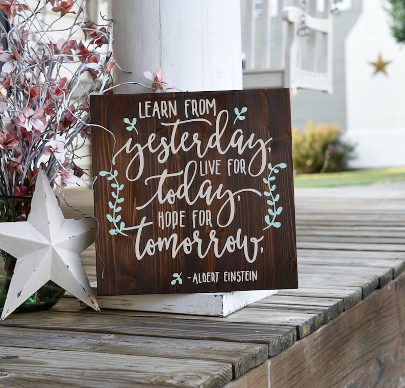 May include: A wooden sign with a dark brown stain and a white painted quote: "Learn from yesterday, live for today, hope for tomorrow." The quote is attributed to Albert Einstein. The sign has a rustic look and is decorated with green leaves.