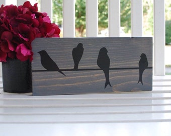 May include: A gray wooden sign with a silhouette of five birds perched on a wire. The birds are black and the wire is white. The sign is hanging on a white picket fence.