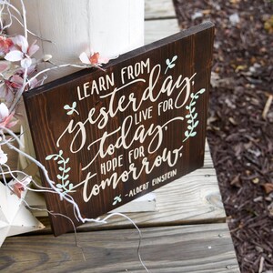 May include: A dark brown wooden sign with a white painted quote: "Learn from yesterday, live for today, hope for tomorrow." The quote is attributed to Albert Einstein. The sign has a rustic look and is decorated with green leaves.