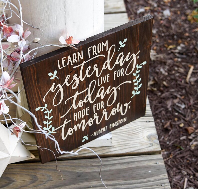 May include: A dark brown wooden sign with a white painted quote: "Learn from yesterday, live for today, hope for tomorrow." The quote is attributed to Albert Einstein. The sign has a rustic look and is decorated with green leaves.
