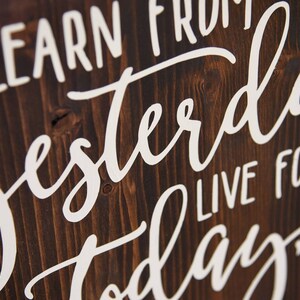 May include: Wooden sign with the text "Learn from yesterday, live for today" written in white paint. The background is dark brown wood with a light blue floral design.