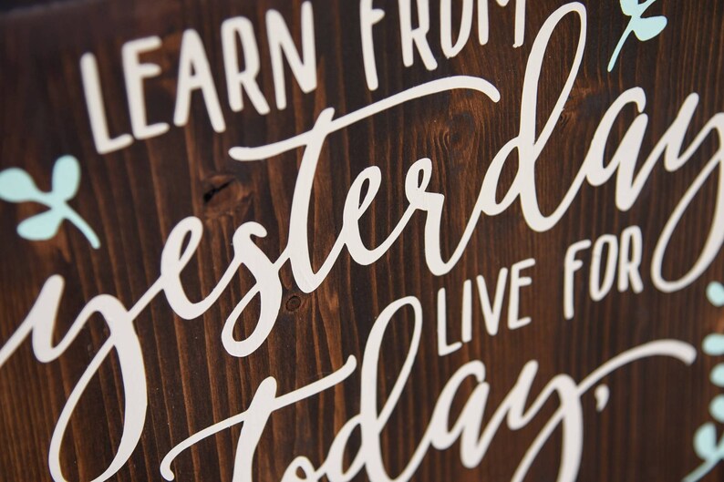 May include: Wooden sign with the text "Learn from yesterday, live for today" written in white paint. The background is dark brown wood with a light blue floral design.