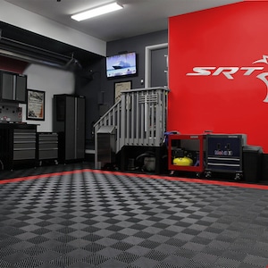 May include: A garage with a red wall featuring the SRT logo and a black and gray checkered floor. The garage has a workbench with toolboxes, a set of stairs, and a television mounted on the wall.