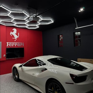 May include: A white Ferrari sports car parked in a garage with a black and red ceiling. The ceiling has a hexagonal pattern of white LED lights. The garage wall has the Ferrari logo and the word "Ferrari" in white letters.