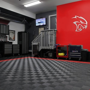 May include: A garage with a red wall featuring a silver Dodge Challenger logo. The floor is covered in black and gray interlocking tiles with a red border. There are various tools and storage units throughout the garage.
