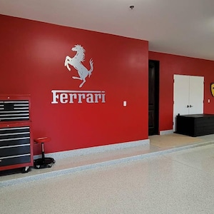 May include: A red garage with a white Ferrari logo on the wall. The logo features a rearing horse with the word "Ferrari" underneath. There is a red toolbox with black drawers in the foreground.