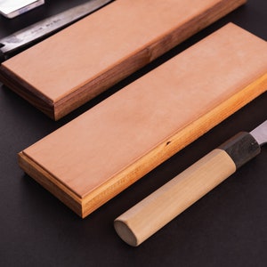 Puede incluir: Dos piedras de afilar de madera con superficies de cuero para afilar cuchillos. Las piedras están sobre una superficie negra con un cuchillo de mango de madera.