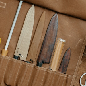 以下が含まれることがあります： 様々なキッチンナイフ、砥石、その他の道具が入った茶色のキャンバス製ナイフロール。ナイフは木製の柄と様々なサイズと色の刃を持っています。黄色のヘラとハサミも写っています。