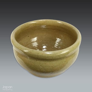 May include: A small, round, ceramic bowl with a light olive green glaze. The bowl has a simple, unadorned design with a slightly textured surface. The interior reflects light, and the bowl appears to be handmade.