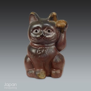 以下が含まれることがあります： 片足を上げた座り姿勢の濃い茶色の陶器製猫の置物。猫は滑らかで光沢のある仕上げで、尖った耳と中央に装飾が付いた小さな首輪が特徴です。目は円形で、ひげは彫り込まれています。