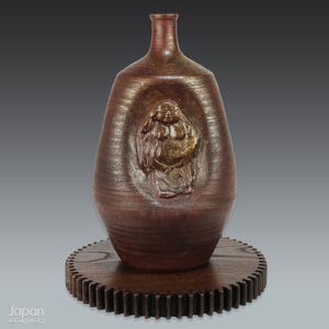 May include: A brown ceramic vase with a textured surface and a relief carving of a Buddha figure. The vase is set on a wooden, gear-shaped base. The vase has a narrow neck and a rounded body.