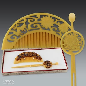 Op de afbeelding: Een verzameling antieke Japanse haaraccessoires, waaronder een grote, ingewikkeld gesneden kam, een kleinere kam, een haarspeld en een decoratieve haarstok. De stukken zijn lichtgeel van kleur met bloemen- en krulontwerpen. De set wordt gepresenteerd in een decoratieve doos.