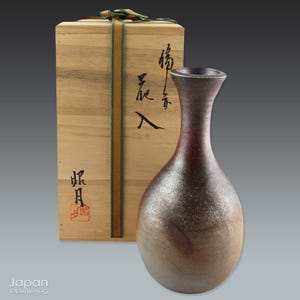 Könnte beinhalten: Eine braune und graue Keramikvase mit einem runden Körper und einem schmalen Hals. Die Vase steht auf einer Holzkiste mit japanischen Schriftzeichen.