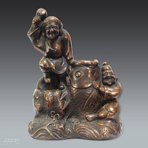 May include: A bronze sculpture depicting two figures with a large fish. One figure is seated and holding a mallet, while the other embraces the fish. The sculpture has a detailed, aged appearance, with visible textures and a dark brown patina.