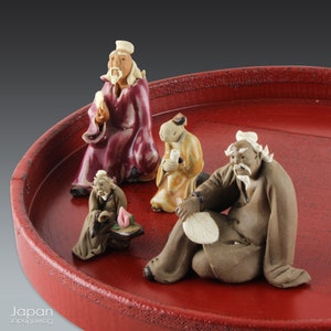 May include: A set of four small ceramic figurines of people, each with a different expression and pose, sitting on a red tray. The figurines are made in a traditional style and are painted in muted colors.