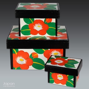 May include: Three nesting boxes with black lids and white bases decorated with red camellia flowers and green leaves.