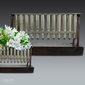 Op de afbeelding: Een vintage houten en metalen standaard met een reeks metalen buizen. De standaard wordt gebruikt om bloemen of andere decoratieve items te houden. De standaard is gemaakt van donker hout en heeft een rustieke uitstraling.