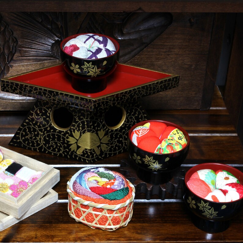 May include: A set of five traditional Japanese fabric covered bowls, known as "tsumami zaiku", with colorful floral patterns. The bowls are arranged on a black and red wooden stand with intricate carvings.