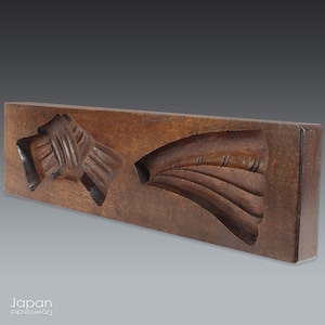 May include: A wooden mold for making Japanese confectionery, featuring two designs: a bow and a fan. The mold is dark brown and has a smooth, polished surface.