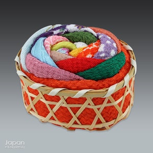 May include: A small, round, woven basket with a lid. The lid is made of colorful fabric, with a variety of patterns and colors, including pink, blue, green, yellow, and red. The basket is made of natural materials and has a traditional Japanese design.