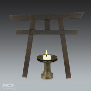 May include: A weathered, brown metal torii gate stands behind a small, antique candle holder with a lit tea light. The image has a gradient grey background. The word "Japan" is in the bottom left corner.