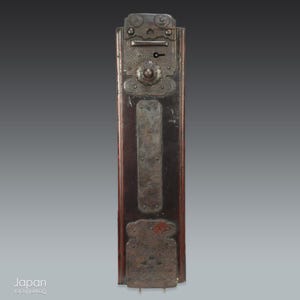 May include: An antique Japanese tansu door with ornate metal hardware. The door features a dark wood frame with decorative metal plates, a keyhole, and intricate floral and geometric designs. The metal has a weathered, aged appearance.