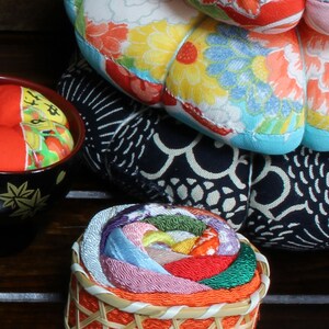May include: A colorful, multi-layered fabric rose pincushion in a woven basket. The pincushion is made of various shades of pink, red, green, blue, and yellow fabric. The basket is orange and has a woven pattern.