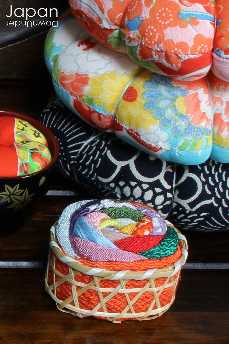 May include: A colorful, multi-layered fabric rose pincushion in a woven basket. The pincushion is made of various shades of pink, red, green, blue, and yellow fabric. The basket is orange and has a woven pattern.
