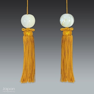 May include: Two white jade beads with gold tassels hanging from a string. The tassels are made of a fine, golden thread and have a decorative knot at the top.