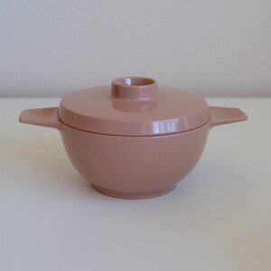 May include: A vintage, light brown, lidded serving bowl. The bowl has a round shape with a flat base and two side handles. The lid has a small, round handle. The bowl is made of a smooth, solid material.