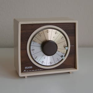 May include: A vintage Sears Kenmore kitchen timer. The timer has a cream-colored plastic frame with a dark wood-grain face. The timer dial is silver with black numbers and a dark brown knob. The Sears Kenmore logo is printed on the bottom.