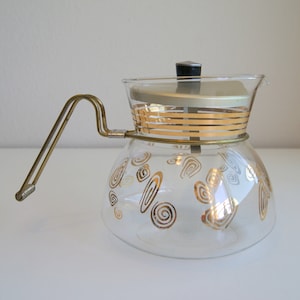 May include: A clear glass coffee pot with a gold metal handle and lid. The pot is decorated with gold spiral and stripe designs. The lid has a black knob. A vintage kitchenware item.
