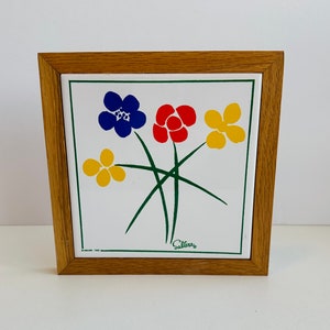 Pode incluir: Um azulejo emoldurado com um design floral. A obra de arte apresenta um buquê de flores estilizadas em azul, vermelho e amarelo, com caules e folhas verdes, sobre um fundo branco. A moldura é feita de madeira castanha clara.
