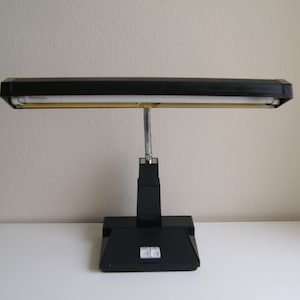 Op de afbeelding: Een zwarte bureaulamp met een rechthoekige kop en een flexibele, zilverkleurige nek. De lamp heeft een rechthoekige basis met twee witte knoppen. De lamp is ontworpen voor gebruik op een bureau en biedt gerichte verlichting.