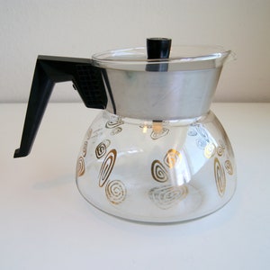 May include: A clear glass coffee pot with a stainless steel top and black handle. The glass carafe is decorated with gold swirl designs. The lid is black. The coffee pot is designed for brewing coffee.