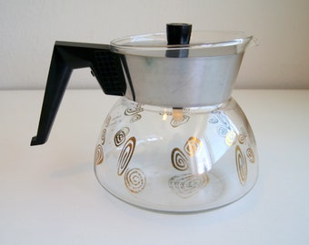 Op de afbeelding: Een glazen koffiepot met een roestvrijstalen bovenkant en een zwarte handgreep. De glazen karaf is versierd met gouden wervelontwerpen. De deksel is zwart. De koffiepot is ontworpen voor het zetten van koffie.