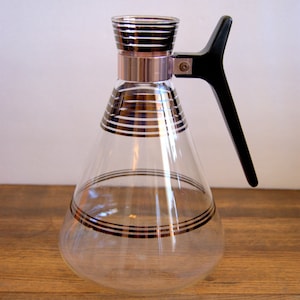 Puede incluir: Una jarra de vidrio transparente con un asa negra y bandas decorativas negras y doradas. La jarra tiene forma cónica y una banda plateada alrededor del cuello. El asa es negra y angular. Un artículo de cocina vintage.