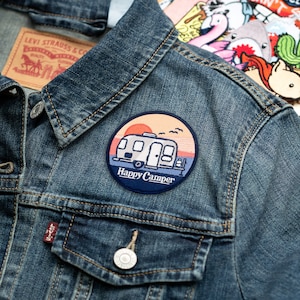 May include: A blue denim jacket with a round embroidered patch featuring a camper van, a sunset, and the text "Happy Camper".