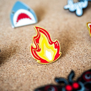 May include: A gold-colored enamel pin shaped like a flame. The flame is red with a yellow center and a white tip.