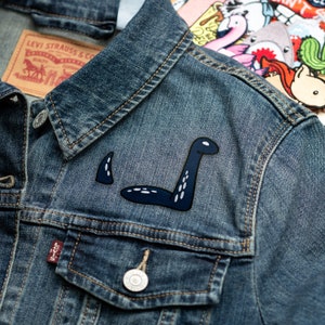 May include: A dark blue denim jacket with a black and white embroidered patch of a sea serpent on the left side of the chest. The jacket has a Levi's label on the left pocket.