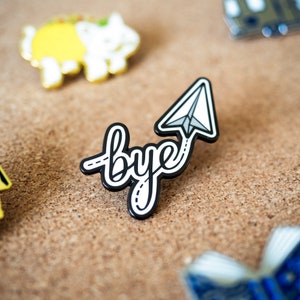 May include: Black enamel pin with the word "bye" in a white cursive font and a white paper airplane with a black outline.