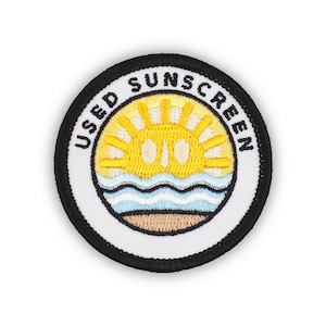 May include: A white circular patch with black stitching around the edge. The patch has a yellow sun with a white center and blue waves at the bottom. The text "USED SUNSCREEN" is written in black around the sun and waves.