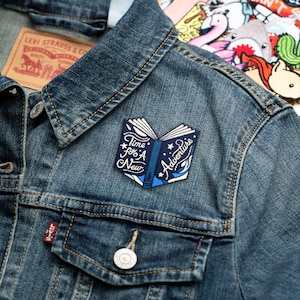 May include: A blue and white embroidered iron-on patch with an open book design. The book has the text "Time for a New Adventure" on it.