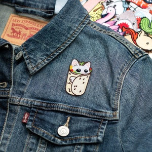 May include: A white cat is wrapped in a burrito. The cat is looking out of the burrito with big eyes. The burrito is on a blue denim jacket.