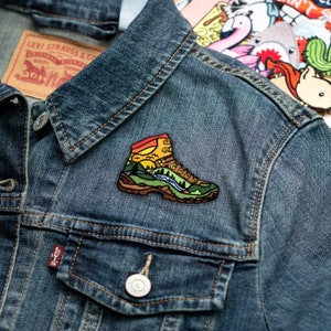 May include: A colourful embroidered patch of a hiking boot with a mountain scene on a denim jacket.
