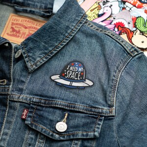 May include: A denim jacket with a patch that reads "I NEED MY SPACE" in white text. The patch is shaped like a UFO with a blue and white design. A Levi Strauss & Co. tag is visible.